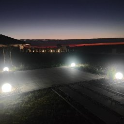 VOLT MASTERS Ernest Wojdyła - Ogród po zmroku z kulistymi lampami oświetlającymi ścieżkę i taras. W tle widoczny budynek i linia horyzontu z delikatną poświatą zachodzącego słońca.