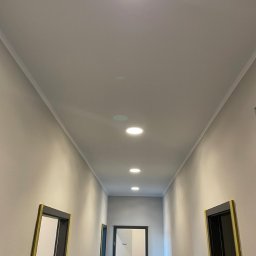 VOLT MASTERS Ernest Wojdyła - Długi korytarz z rzędem równomiernie rozmieszczonych, okrągłych lamp sufitowych. Ściany w odcieniach szarości, widoczne ościeżnice drzwi. Minimalistyczny design.