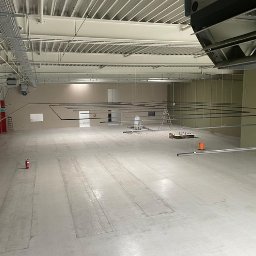 VOLT MASTERS Ernest Wojdyła - Przestronne wnętrze hali z widoczną instalacją podwieszaną, drabina i narzędzia sugerują montaż oświetlenia. Widok z góry na surową podłogę i czerwone słupy.