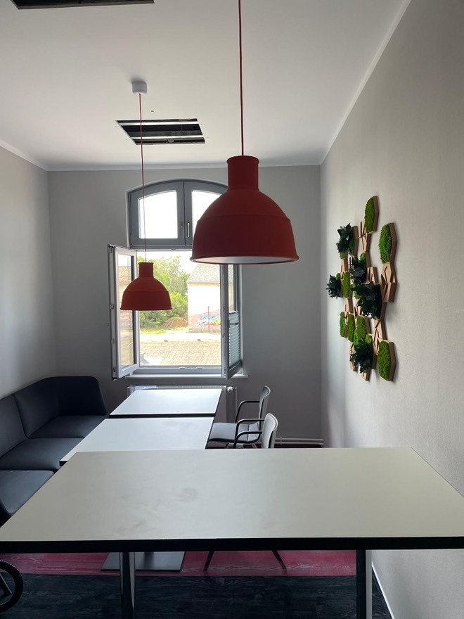 Wnętrze biura z dwoma czerwonymi lampami wiszącymi i oknem z widokiem na zewnątrz. Na ścianie dekoracja z mchu. Widoczny fragment sufitu z otworem.