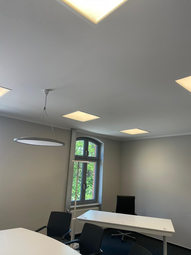 Nowoczesne biuro z oknem: okrągła lampa wisząca, kwadratowe panele LED w suficie, białe biurka i czarne krzesła biurowe. Widok z dołu, minimalistyczny design.