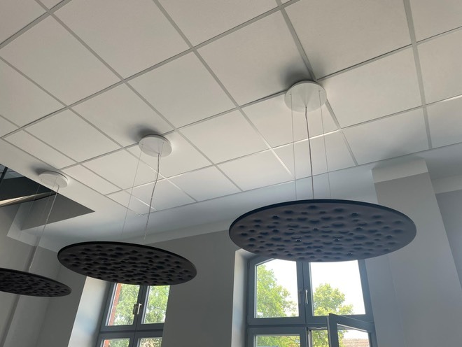 Nowoczesne, okrągłe lampy wiszące z czarnymi, perforowanymi kloszami zamontowane pod kasetonowym sufitem w jasnym pomieszczeniu biurowym.