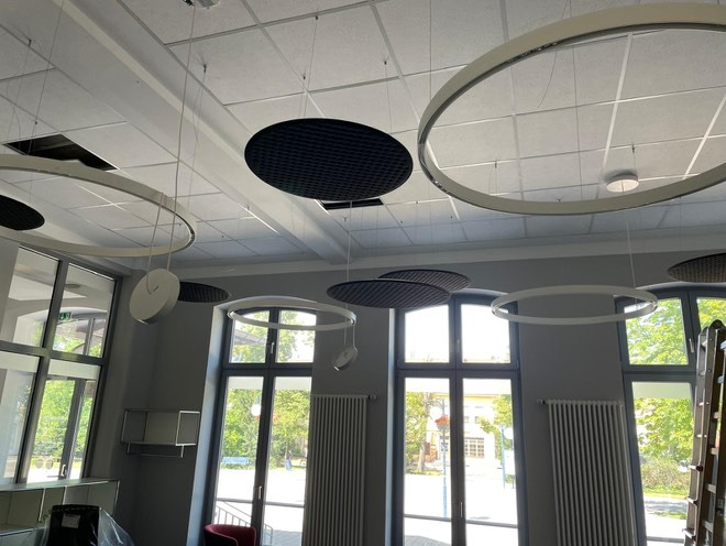 Nowoczesne oświetlenie sufitowe: okrągłe lampy LED i panele akustyczne w jasnym wnętrzu z dużymi oknami. Widoczne niedokończone elementy sufitu podwieszanego.