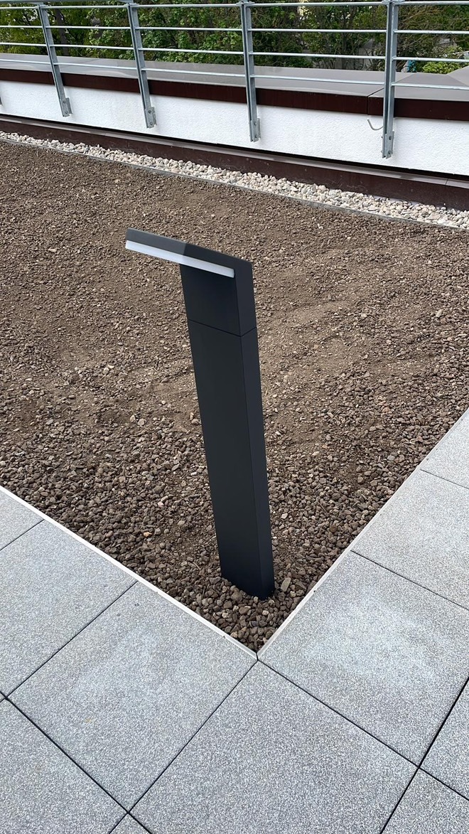 Nowoczesna lampa ogrodowa w kolorze antracytu, oświetlająca rabatę z drobnym żwirem na tle szarej kostki brukowej i metalowej balustrady.