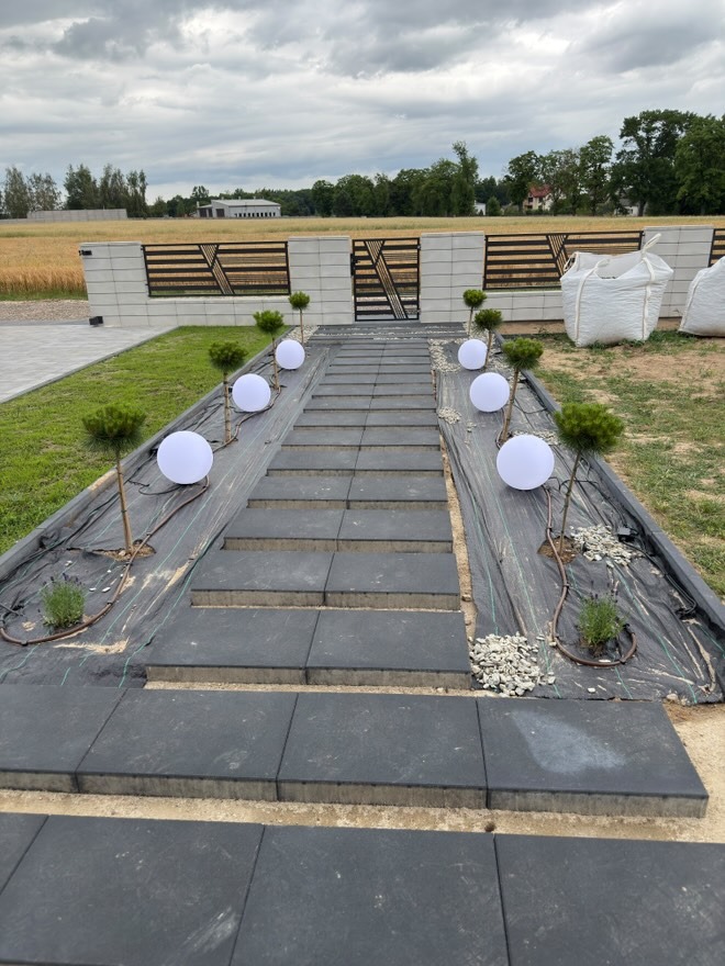 Oświetlona ścieżka z szarych płyt prowadzi do nowoczesnej bramy ogrodowej. Dekoracyjne kule świetlne i drzewka dodają uroku. Widoczne worki z materiałem budowlanym.