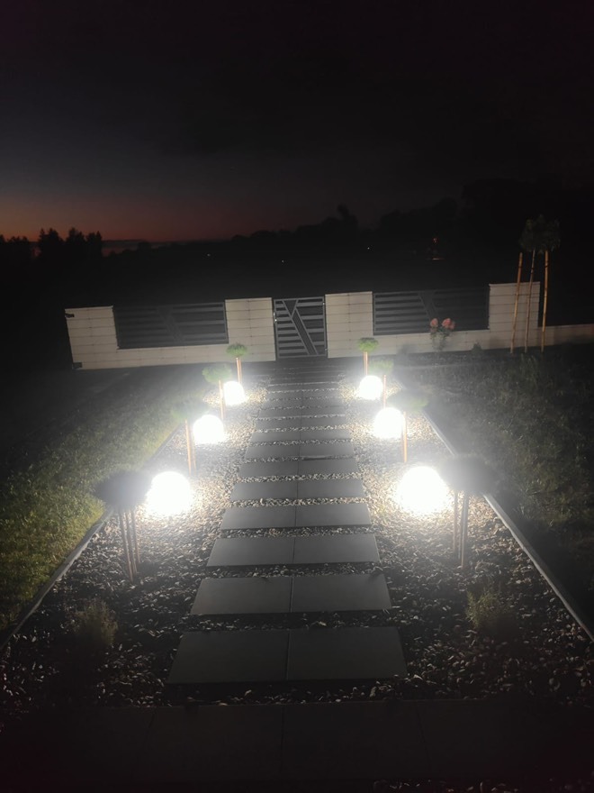 Oświetlona ścieżka z kamieni i żwiru prowadząca do bramki w ogrodzeniu, oświetlona kulistymi lampami ogrodowymi w ciepłym odcieniu, nocne ujęcie.