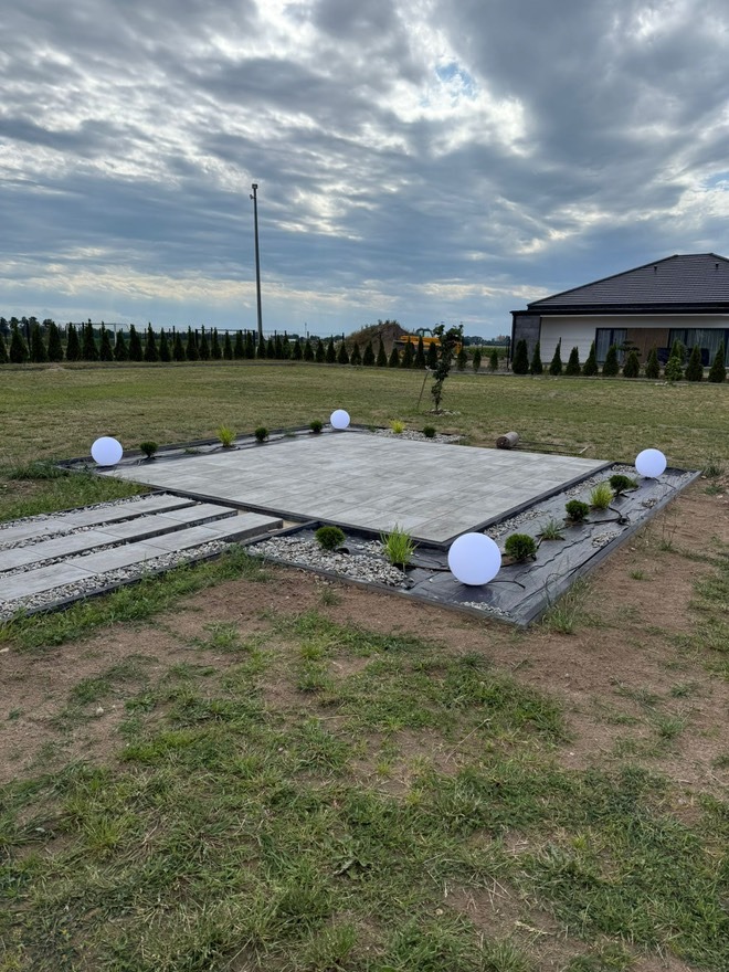 Ogród z podświetlanym tarasem z kostki brukowej, oświetlony kulistymi lampami. W tle widoczny dom i rząd drzew iglastych.