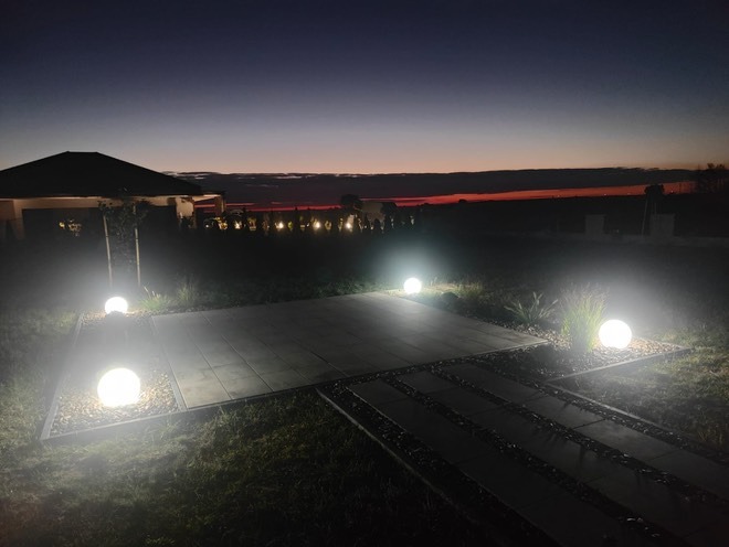 Ogród po zmroku z kulistymi lampami oświetlającymi ścieżkę i taras. W tle widoczny budynek i linia horyzontu z delikatną poświatą zachodzącego słońca.