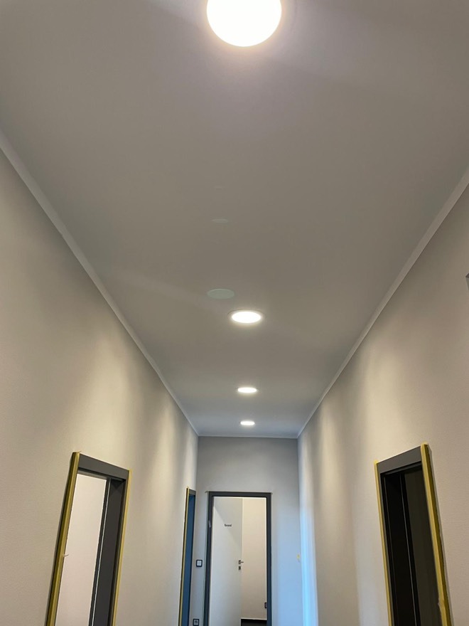 Długi korytarz z rzędem równomiernie rozmieszczonych, okrągłych lamp sufitowych. Ściany w odcieniach szarości, widoczne ościeżnice drzwi. Minimalistyczny design.