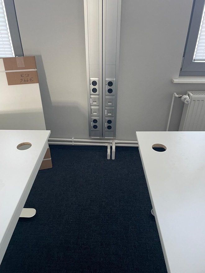 Dwa aluminiowe słupy z gniazdkami elektrycznymi i portami sieciowymi, zamontowane na ścianie w biurze, widoczne białe biurka i okno z żaluzjami.