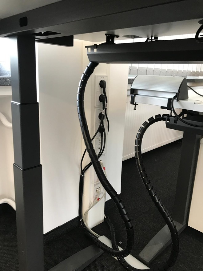 Elektryczne gniazdka pod biurkiem z czarnymi kablami w spiralnych osłonach, zamontowane na białej ściance. Widok z dołu na instalację.