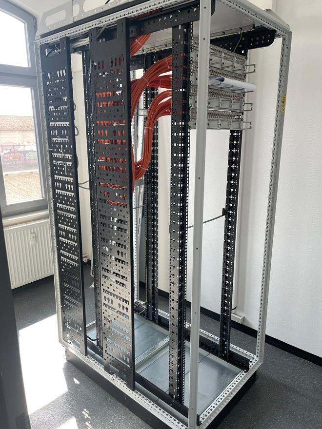 Metalowa szafa rack z pomarańczowymi kablami, widoczne panele krosownicze i okablowanie strukturalne wewnątrz, oświetlone światłem dziennym z okna.