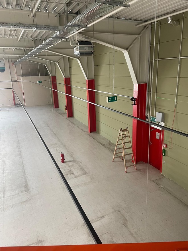 Wysoka hala z instalacjami pod sufitem, czerwonymi słupami i znakami ewakuacyjnymi. Widoczne kable, koryta kablowe, drabina i gaśnice.