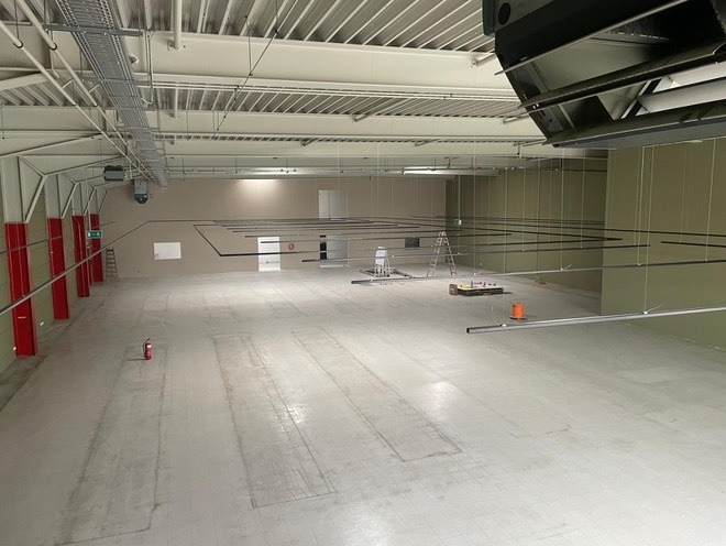 Przestronne wnętrze hali z widoczną instalacją podwieszaną, drabina i narzędzia sugerują montaż oświetlenia. Widok z góry na surową podłogę i czerwone słupy.