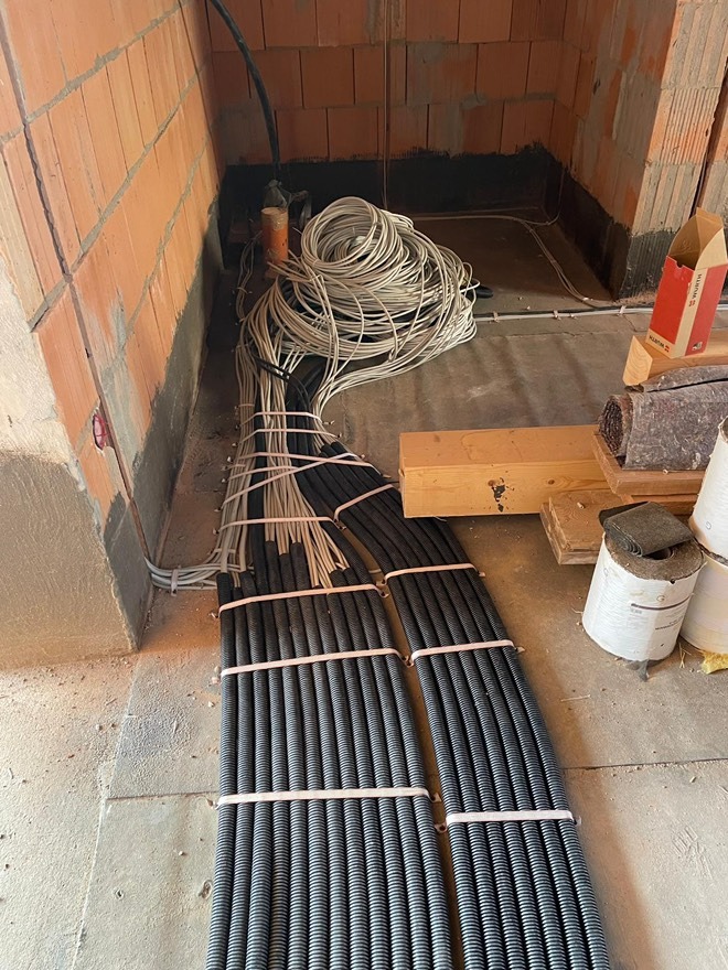 Instalacja elektryczna w surowym stanie: wiązki przewodów w peszlach i luźne kable na podłodze, widoczne ściany z cegły.