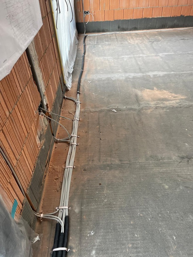 Szare kable elektryczne prowadzone wzdłuż ściany z czerwonej cegły na podłodze w surowym stanie, widok z góry, etap prac instalacyjnych.