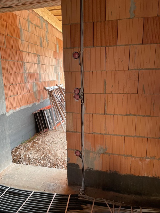 Surowa ściana z cegły z poprowadzonymi przewodami elektrycznymi i puszkami pod gniazdka. Widoczne rury instalacyjne na podłodze i drabina w tle.