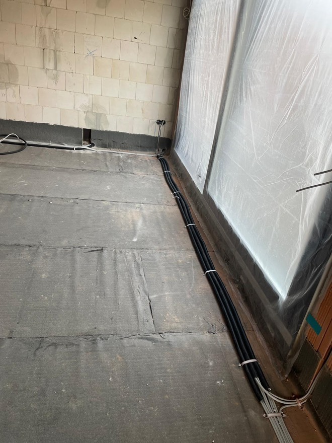 Kable elektryczne ułożone wzdłuż ściany w surowym wnętrzu. Widoczne przewody w peszlu, ściana z białego pustaka i folia ochronna na oknie.