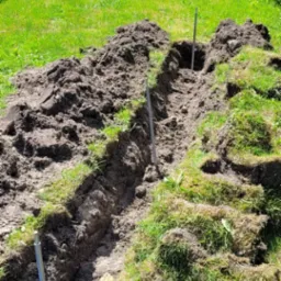 Wykopany rów w trawie z wbitymi metalowymi prętami, prawdopodobnie przygotowanie pod kable. W tle słupy energetyczne i zieleń.