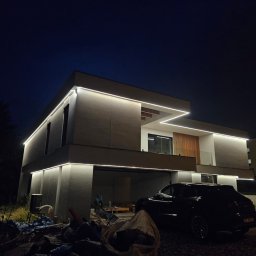 Instalacje Elektryczne KONTAKT - Nowoczesny dom jednorodzinny z podświetleniem LED na elewacji, widoczny nocą, z zaparkowanym samochodem. Minimalistyczny design budynku z oświetleniem liniowym.