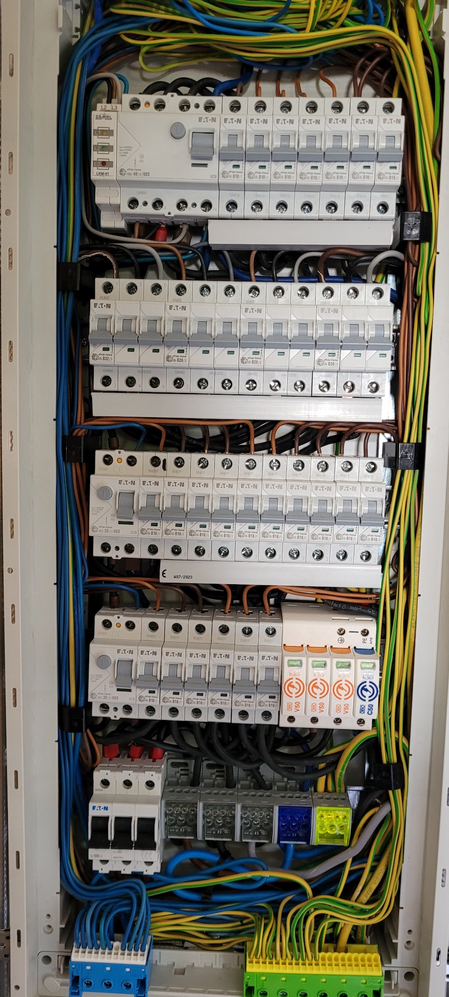 Rozdzielnica elektryczna z licznymi bezpiecznikami EATON i okablowaniem w kolorach niebieskim, żółtym i brązowym. Widok z bliska na zamontowane elementy.