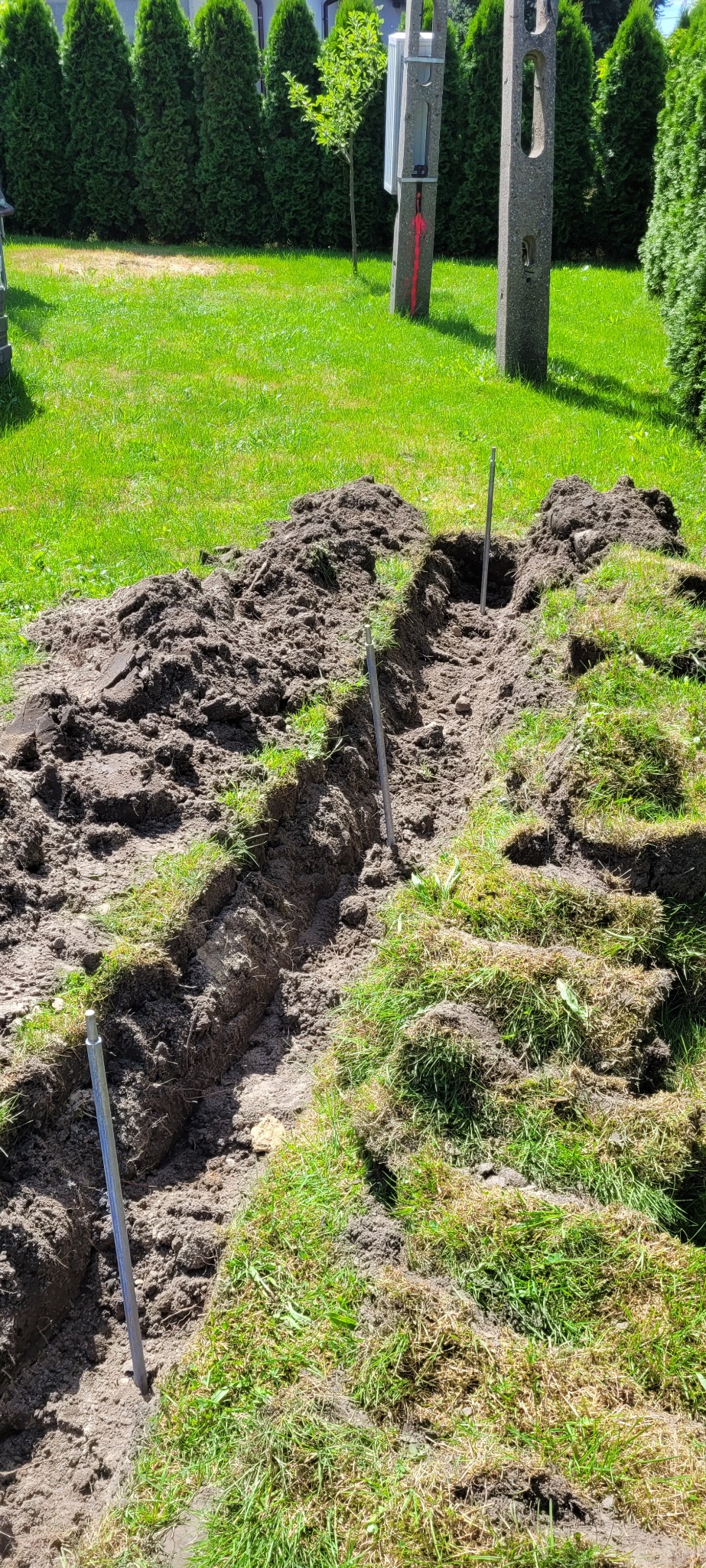 Wykopany rów w trawie z wbitymi metalowymi prętami, prawdopodobnie przygotowanie pod kable. W tle słupy energetyczne i zieleń.