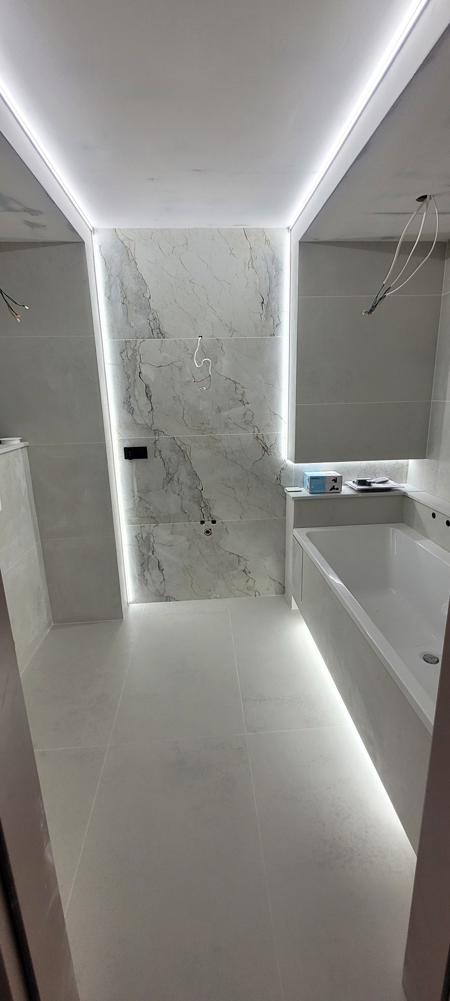 Nowoczesna łazienka z podświetleniem LED: sufit, ściana z marmurowymi płytkami i wanna. Widoczne przewody elektryczne.