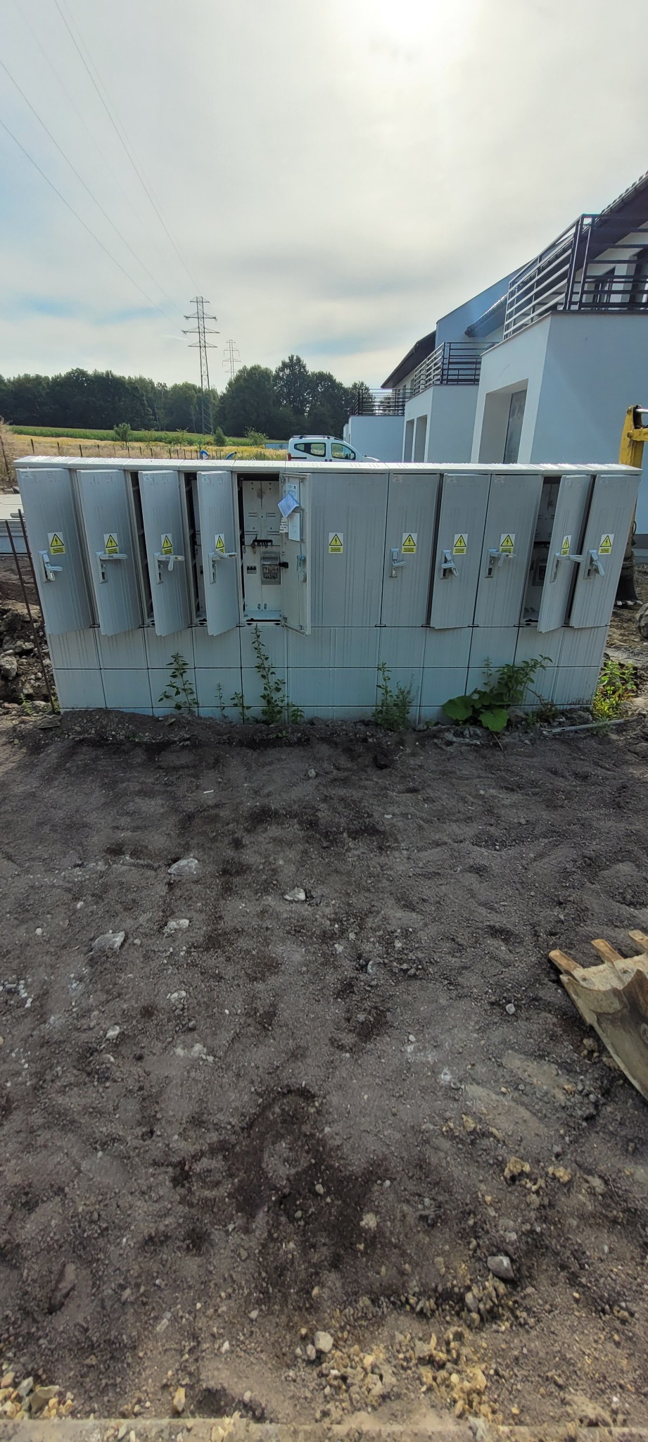 Szara szafa elektryczna z otwartymi drzwiczkami na zewnątrz budynku, z widocznymi elementami instalacji wewnątrz. Na drzwiczkach znaki ostrzegawcze.
