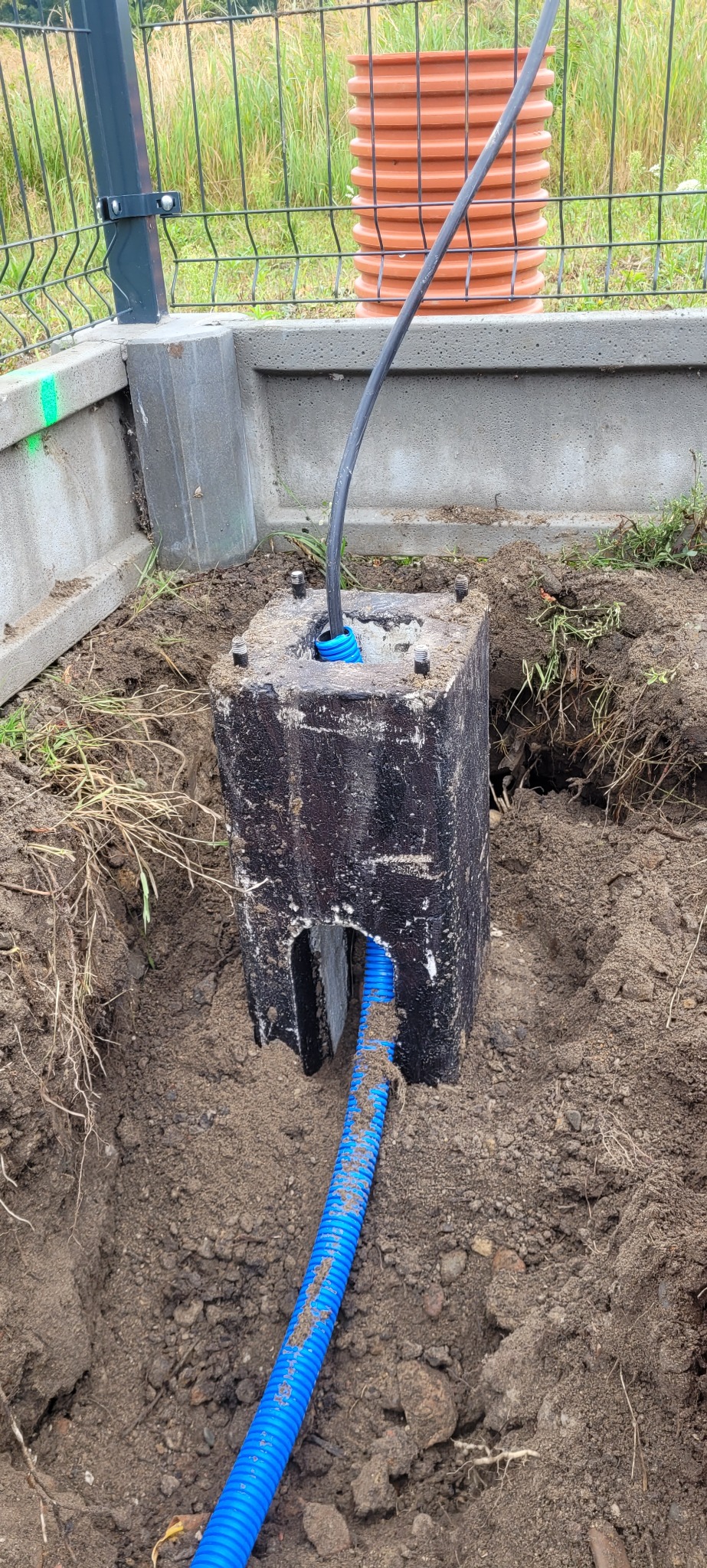 Betonowy fundament z niebieską rurą karbowaną i czarnym kablem, wystający z ziemi, prawdopodobnie element instalacji elektrycznej zewnętrznej.