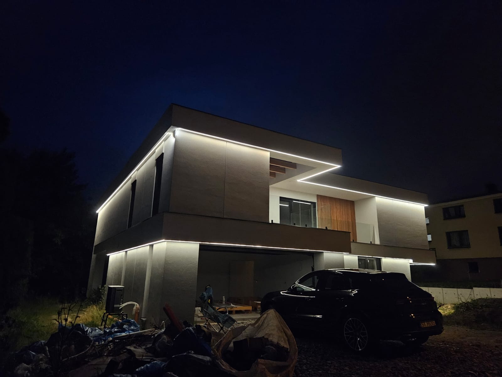 Nowoczesny dom jednorodzinny z podświetleniem LED na elewacji, widoczny nocą, z zaparkowanym samochodem. Minimalistyczny design budynku z oświetleniem liniowym.
