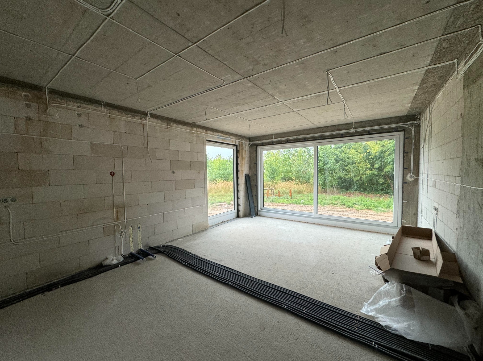 Surowy stan wnętrza z widocznymi przewodami elektrycznymi ułożonymi na podłodze i poprowadzonymi po suficie, okna z widokiem na zieleń.