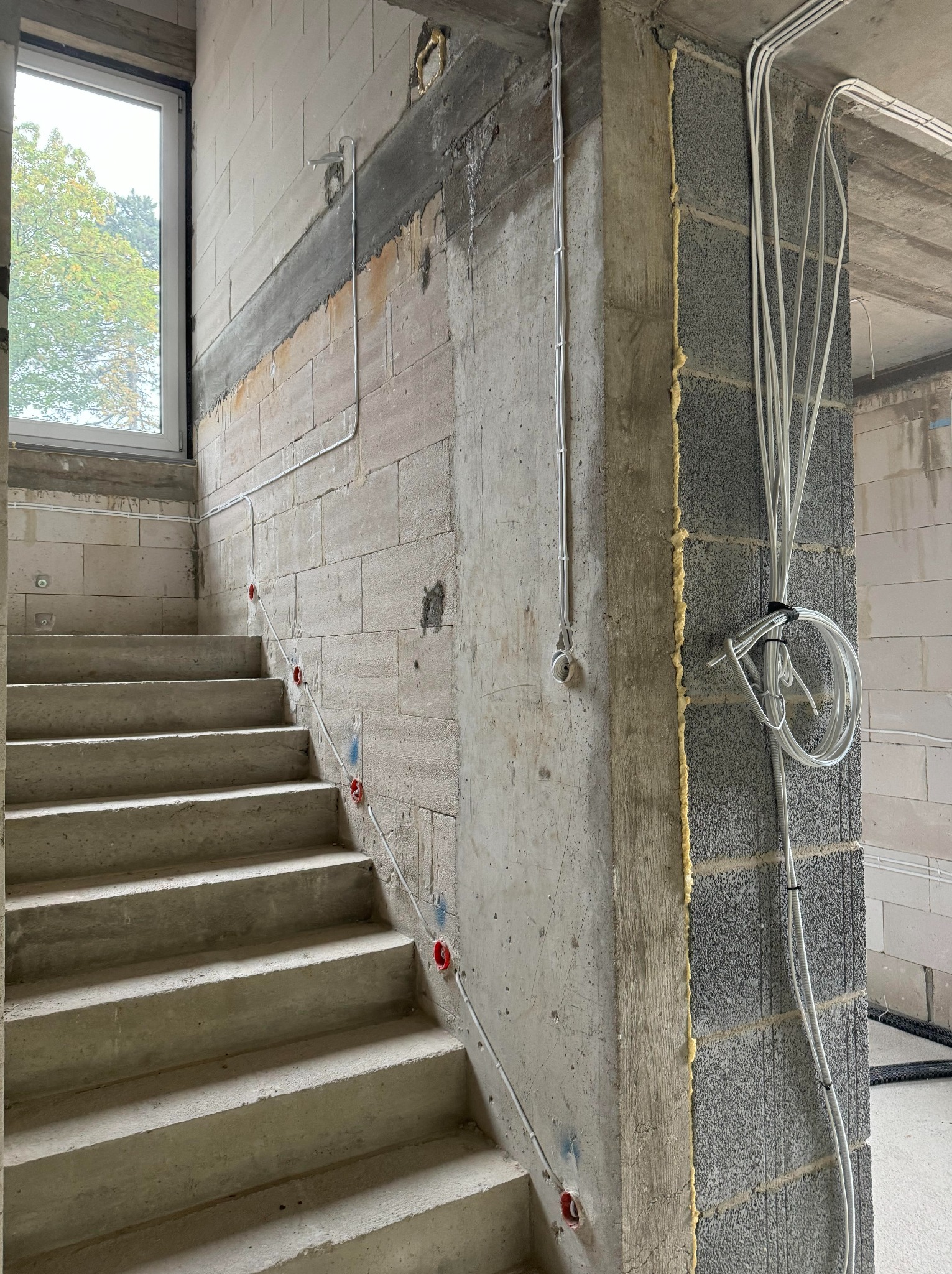 Surowe schody z widoczną instalacją elektryczną w trakcie montażu, przewody prowadzone wzdłuż ściany z bloczków, okno w tle. Wnętrze w stanie deweloperskim.