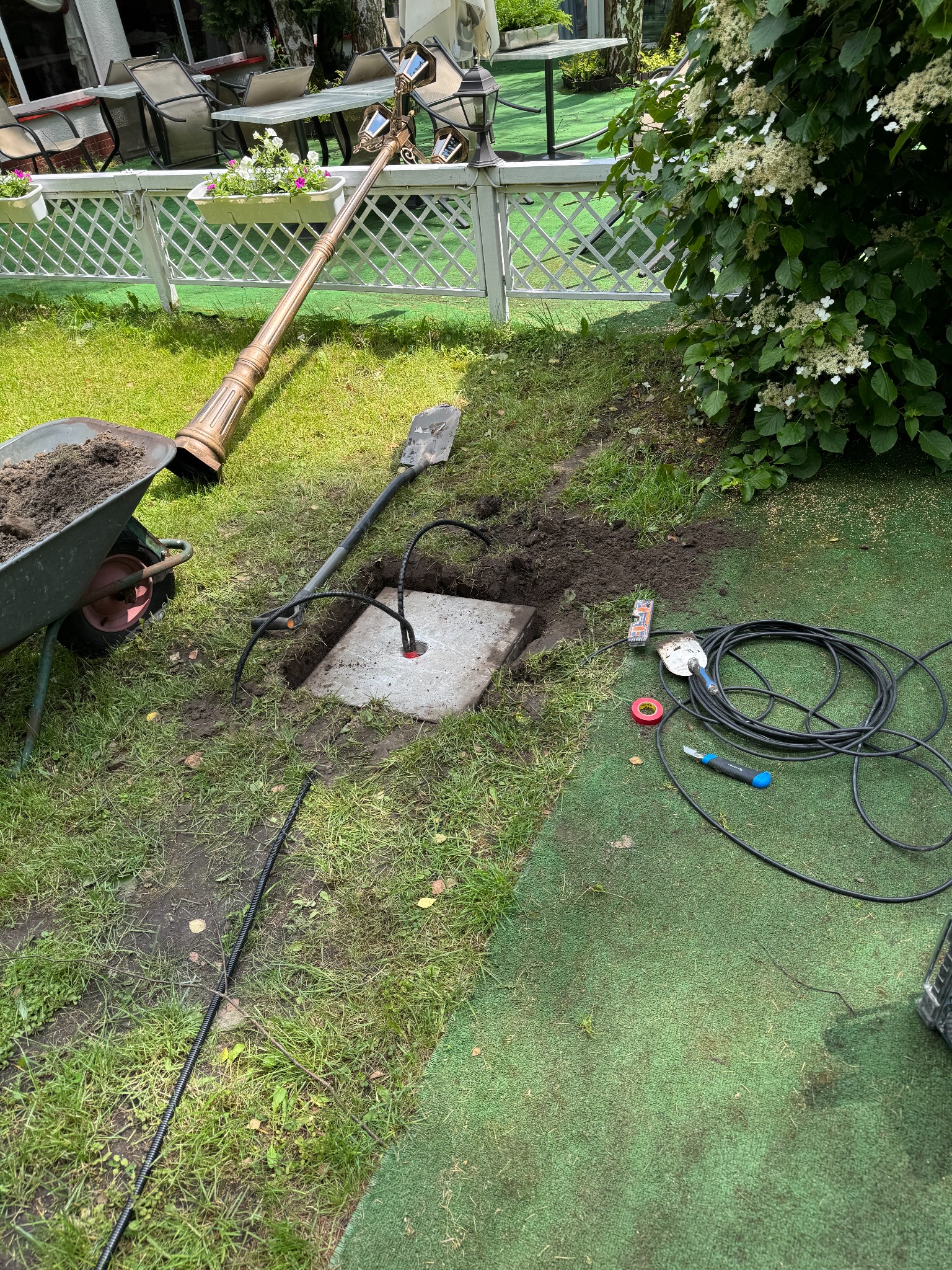 Instalacja oświetlenia ogrodowego: kabel w ziemi, lampa leżąca obok, narzędzia pracy. Widok na trawnik, krzewy i elementy architektury ogrodowej.