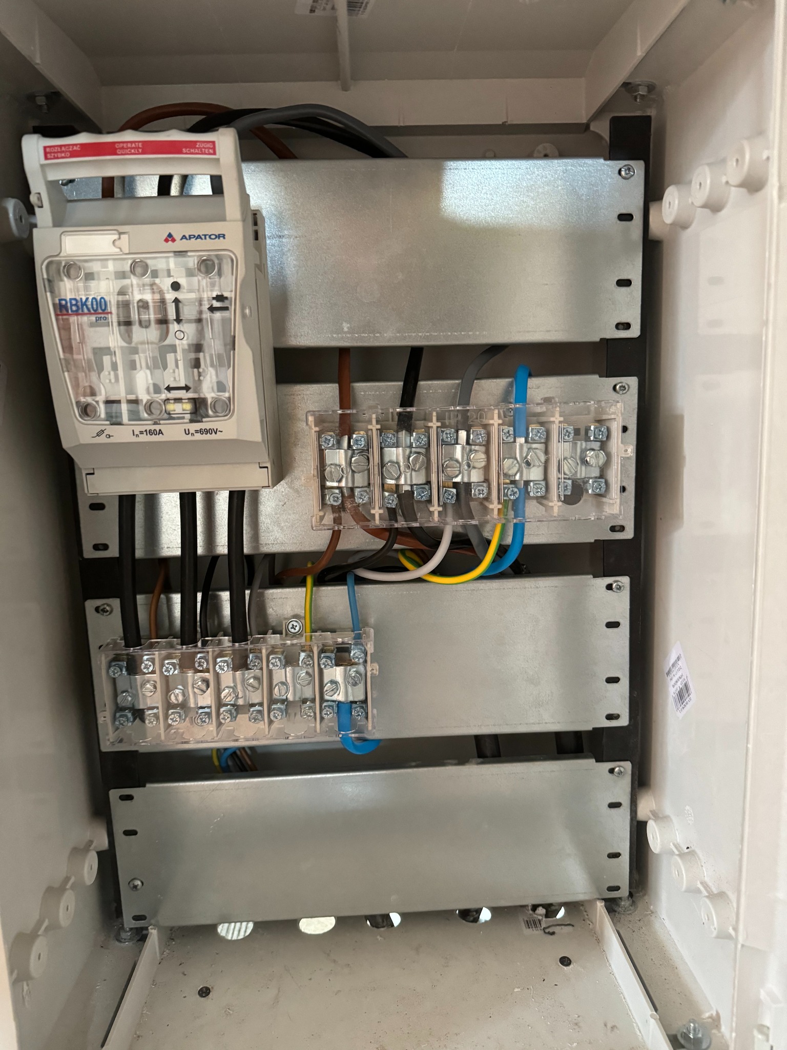 Rozdzielnica elektryczna Apator RBK00 z widocznymi przewodami i złączkami. Instalacja wewnątrz białej obudowy. Widoczne kable w kolorach: czarny, brązowy, niebieski, żółto-zielony.