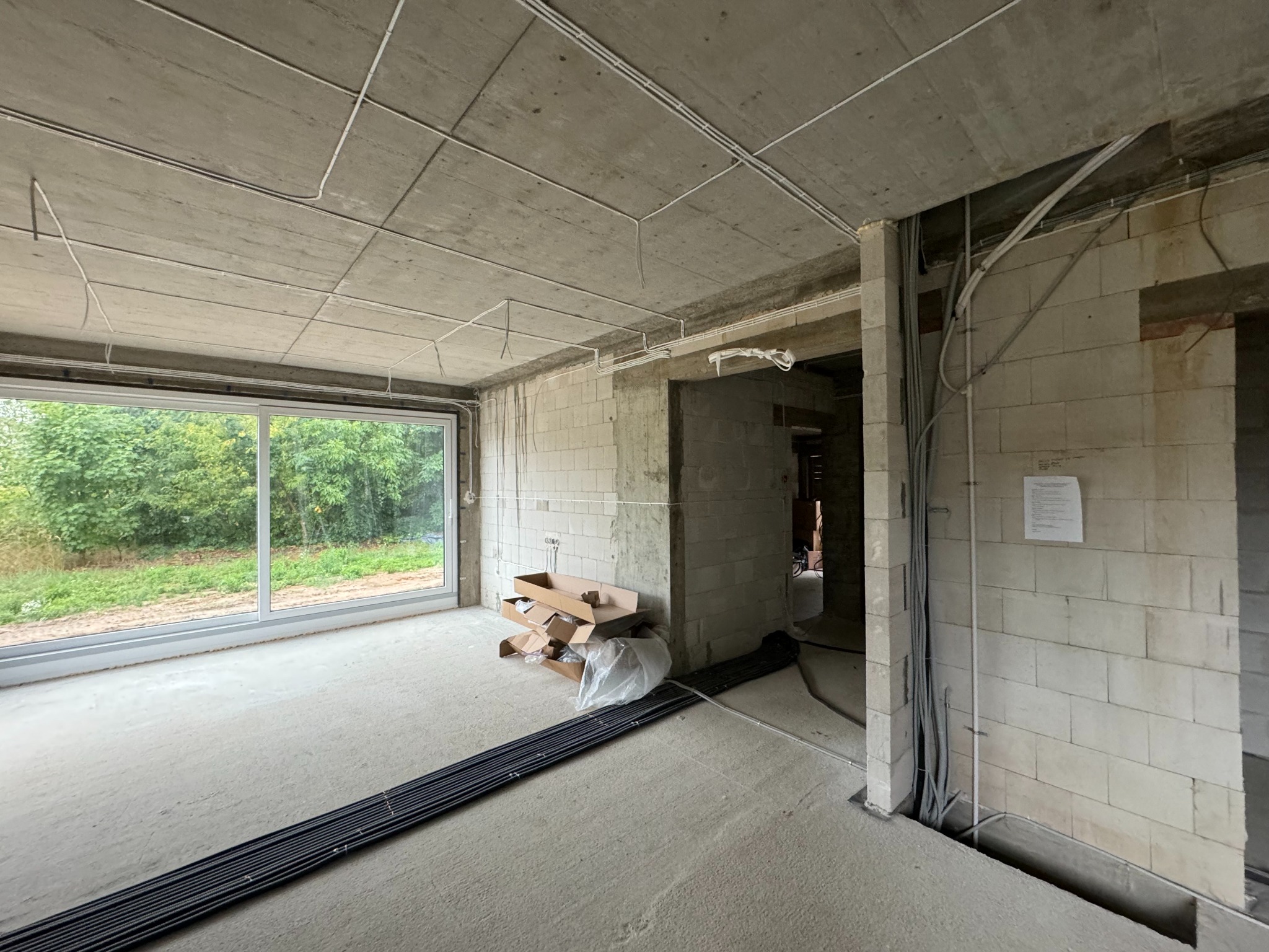 Surowy stan: widok wnętrza w trakcie instalacji elektrycznej. Kable prowadzone po suficie i ścianach, puste przestrzenie, okno z widokiem na zieleń. Układanie kabli w nowym budynku.