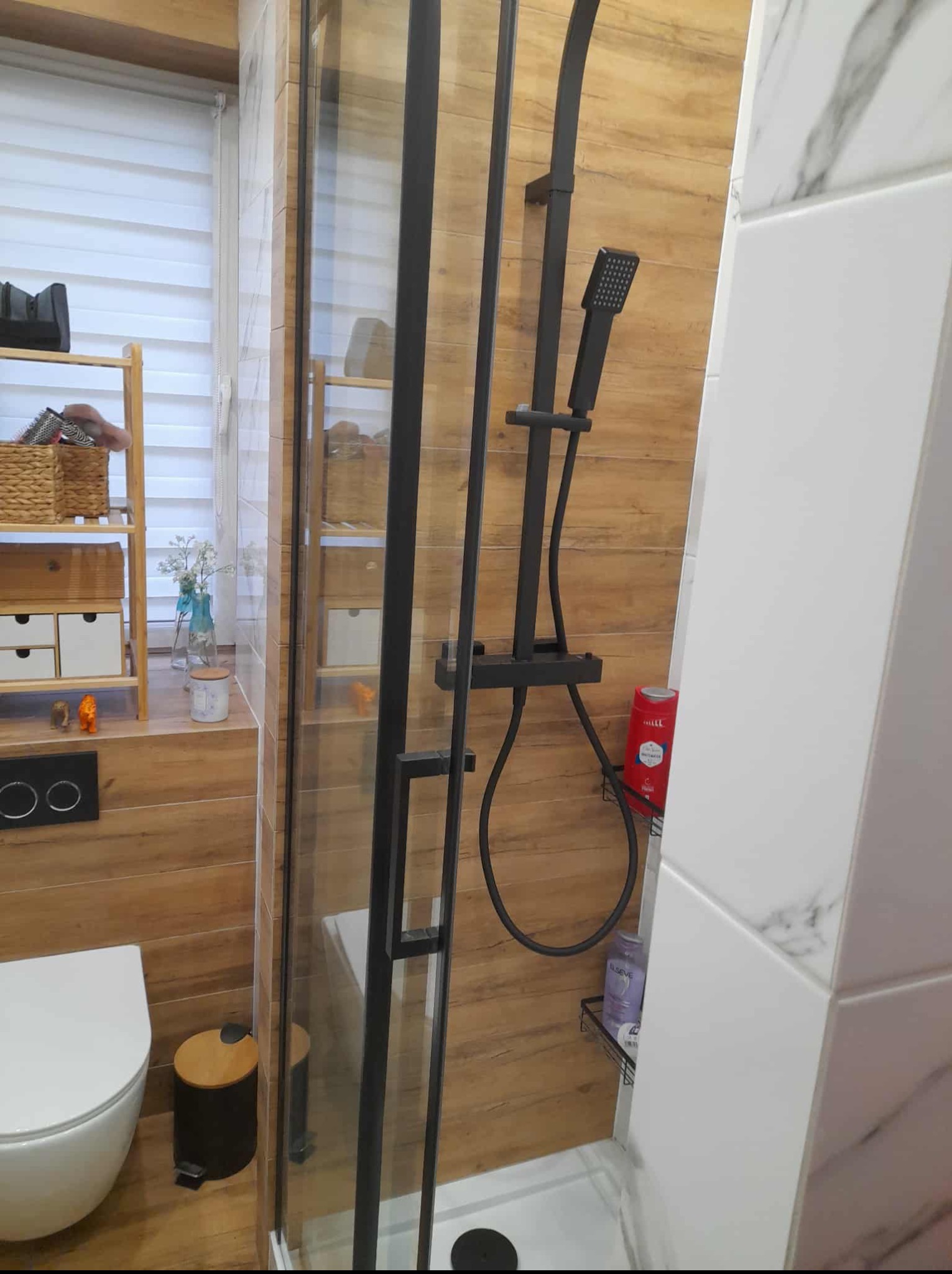 Nowoczesna łazienka z kabiną prysznicową typu walk-in, drewnopodobnymi i marmurowymi płytkami oraz czarną armaturą. Widoczny fragment toalety i półki z dekoracjami.