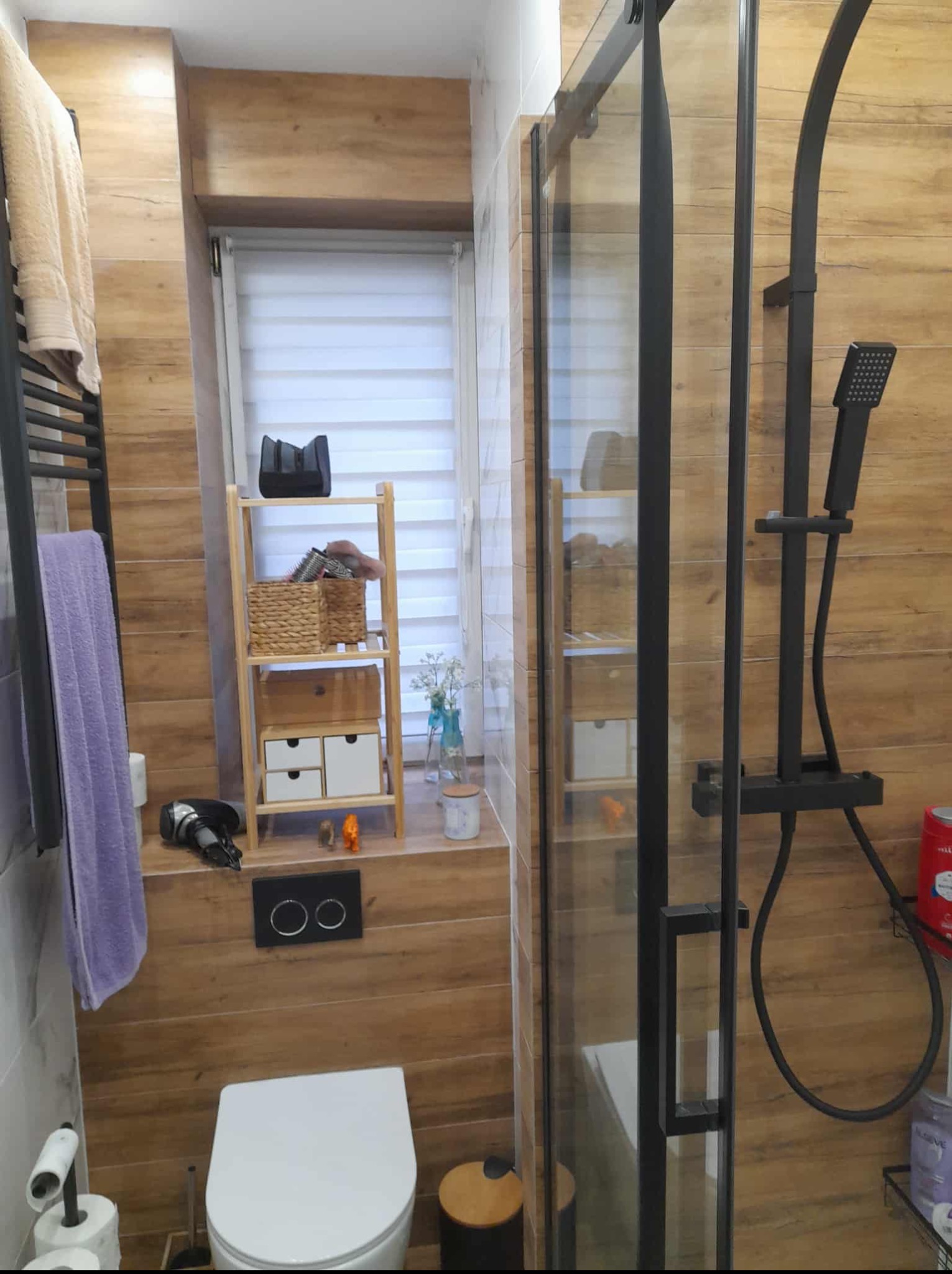 Nowoczesna łazienka z drewnopodobnymi płytkami, czarną armaturą i kabiną prysznicową. Uwagę przykuwa drewniany regał z ozdobnymi koszykami przy oknie.