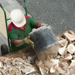 Pracownik w kasku wylewa gruz z wiadra do kontenera. Widoczny wózek transportowy i fragment chodnika z kostki. Ujęcie z góry.