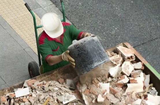 Pracownik w kasku wylewa gruz z wiadra do kontenera. Widoczny wózek transportowy i fragment chodnika z kostki. Ujęcie z góry.