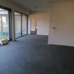 Wnętrze z wylaną, szarą posadzką jastrychową, duże okna tarasowe z widokiem na zewnątrz. Ściany przygotowane do wykończenia, widoczne instalacje elektryczne.