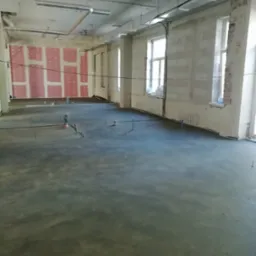 Świeżo wylana, szara posadzka w surowym wnętrzu z instalacjami. Widoczne okna, ściany z bloczków i płyty gipsowe. Detal: rurki wystające z podłoża.