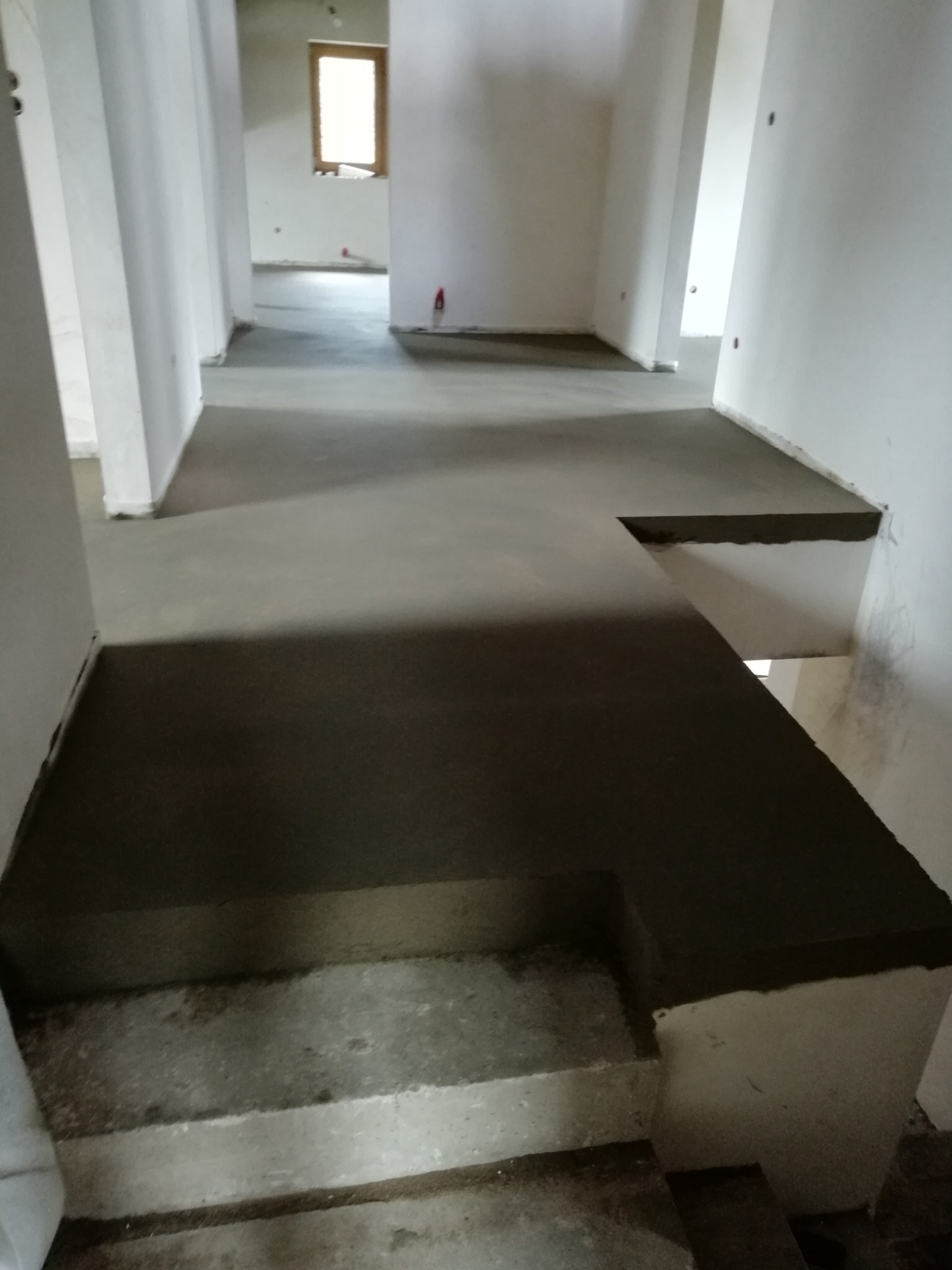 Wnętrze budynku w trakcie prac wykończeniowych. Widoczna wylewka na podłodze i schodach, surowe ściany, okno w oddali. Prace budowlane wewnątrz obiektu.