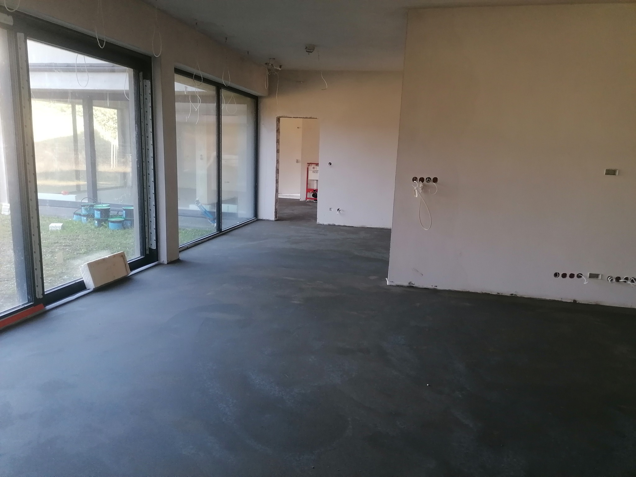Wnętrze z wylaną, szarą posadzką jastrychową, duże okna tarasowe z widokiem na zewnątrz. Ściany przygotowane do wykończenia, widoczne instalacje elektryczne.