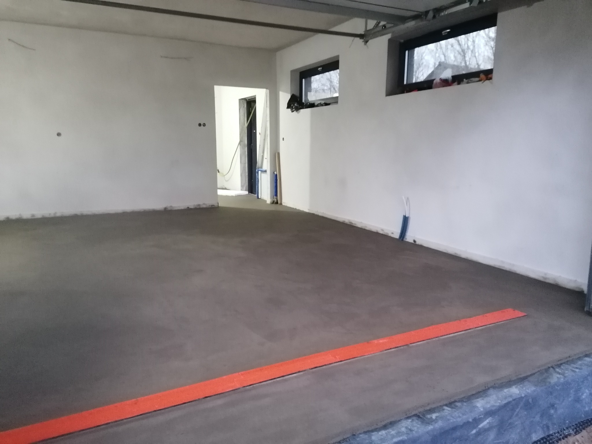 Gładka, szara posadzka jastrychowa w surowym wnętrzu, z widoczną pomarańczową listwą dylatacyjną przy ścianie i oknami w tle. Wnętrze w trakcie wykańczania.
