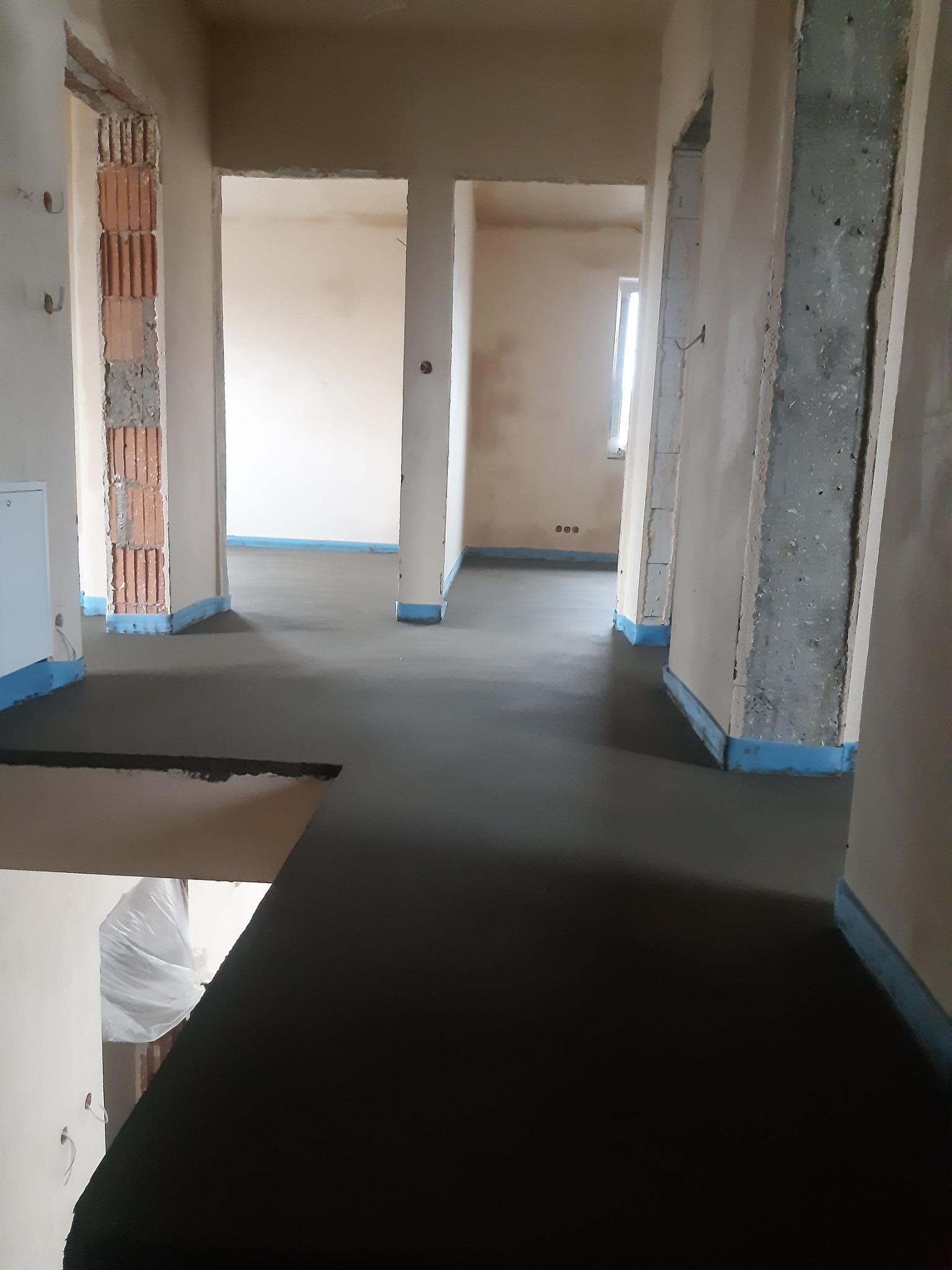 Świeżo wylana, gładka posadzka jastrychowa w jasnym wnętrzu z widocznymi surowymi ścianami i niebieską folią przy krawędziach. Widok z góry.