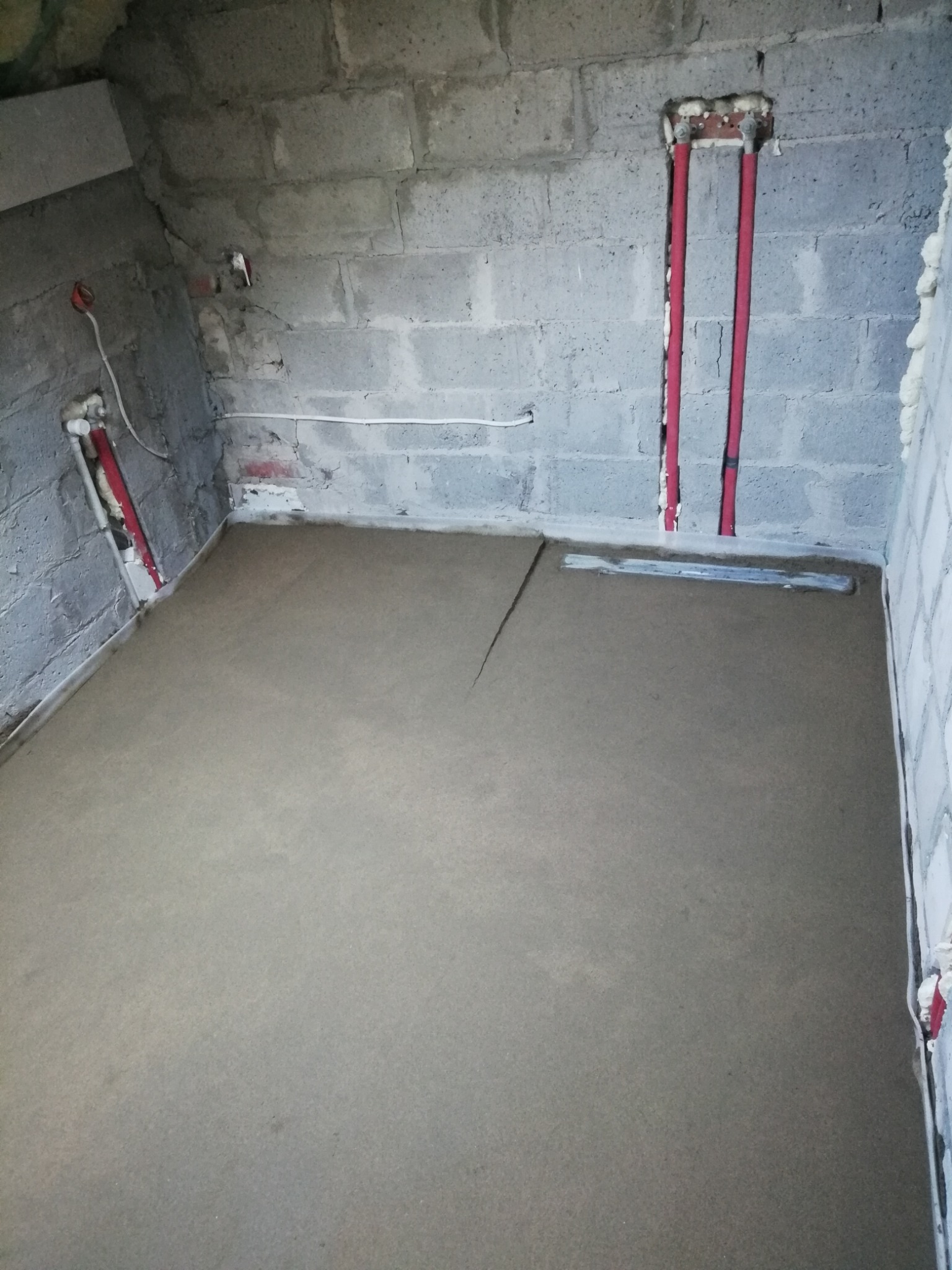Szara, świeżo wylana posadzka jastrychowa w pomieszczeniu z surowymi ścianami z betonu komórkowego. Widoczne czerwone rury instalacji w ścianie i odpływ liniowy.