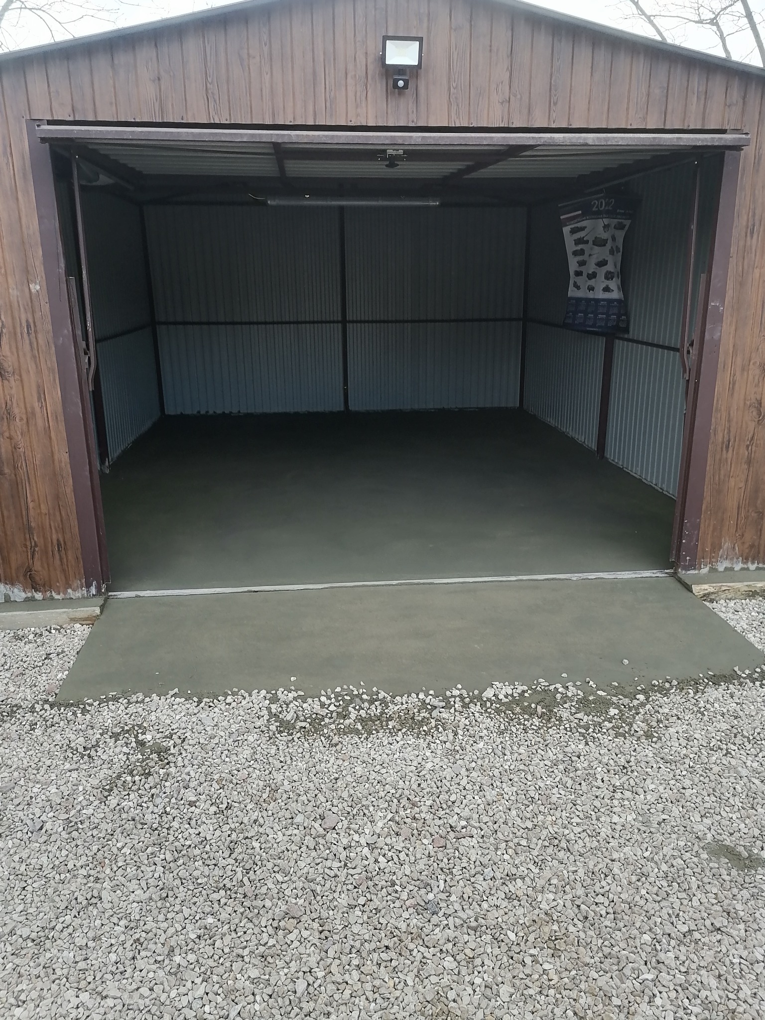 Wnętrze garażu z nową, szarą posadzką. Świeży beton kontrastuje z zewnętrzną, żwirową nawierzchnią. Na ścianie wisi kalendarz.