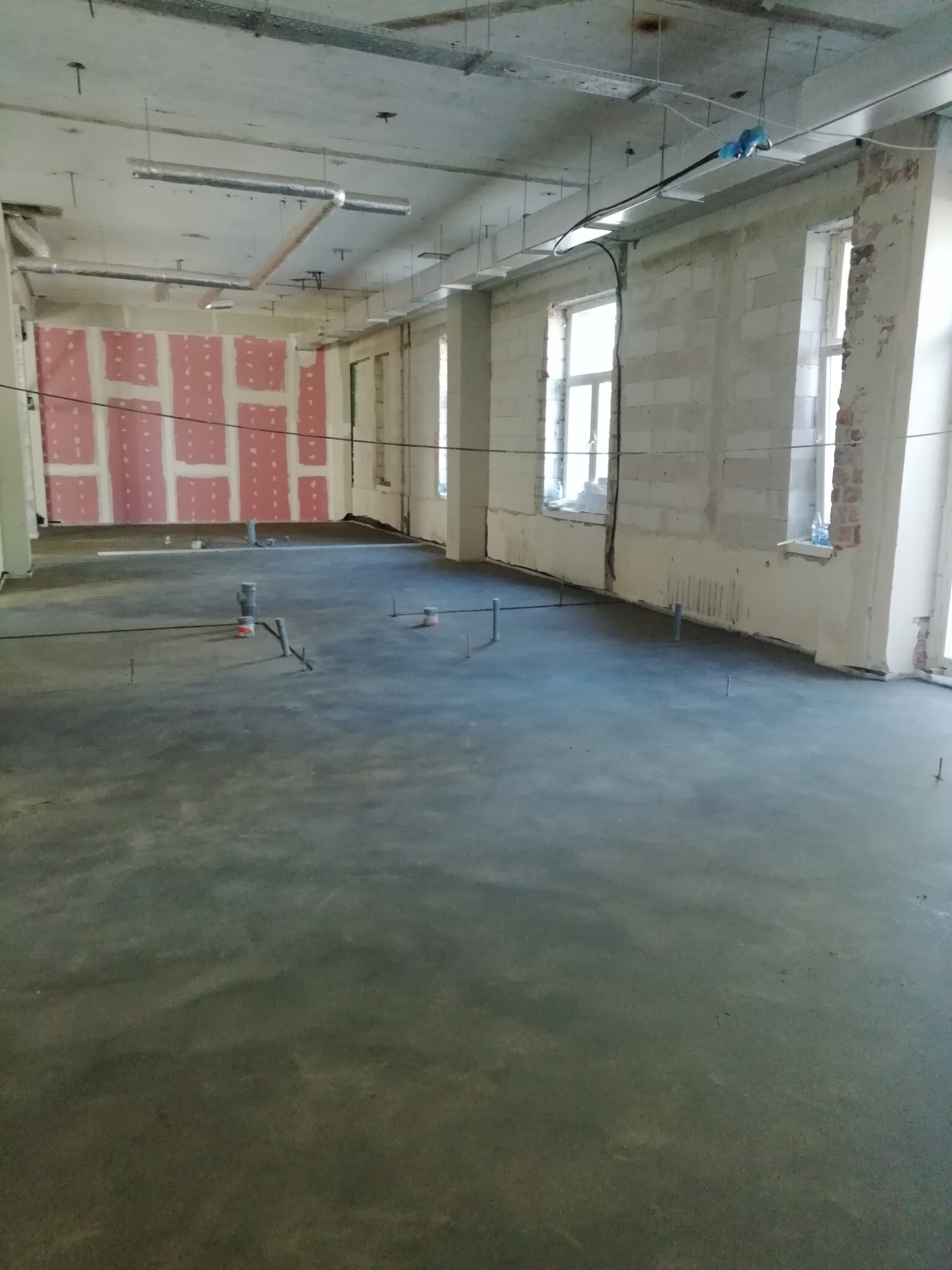Świeżo wylana, szara posadzka w surowym wnętrzu z instalacjami. Widoczne okna, ściany z bloczków i płyty gipsowe. Detal: rurki wystające z podłoża.