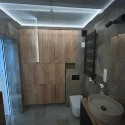 Nowoczesna łazienka z drewnianymi szafkami, szarą armaturą, podświetlanym sufitem i umywalką nablatową. Minimalistyczny design.