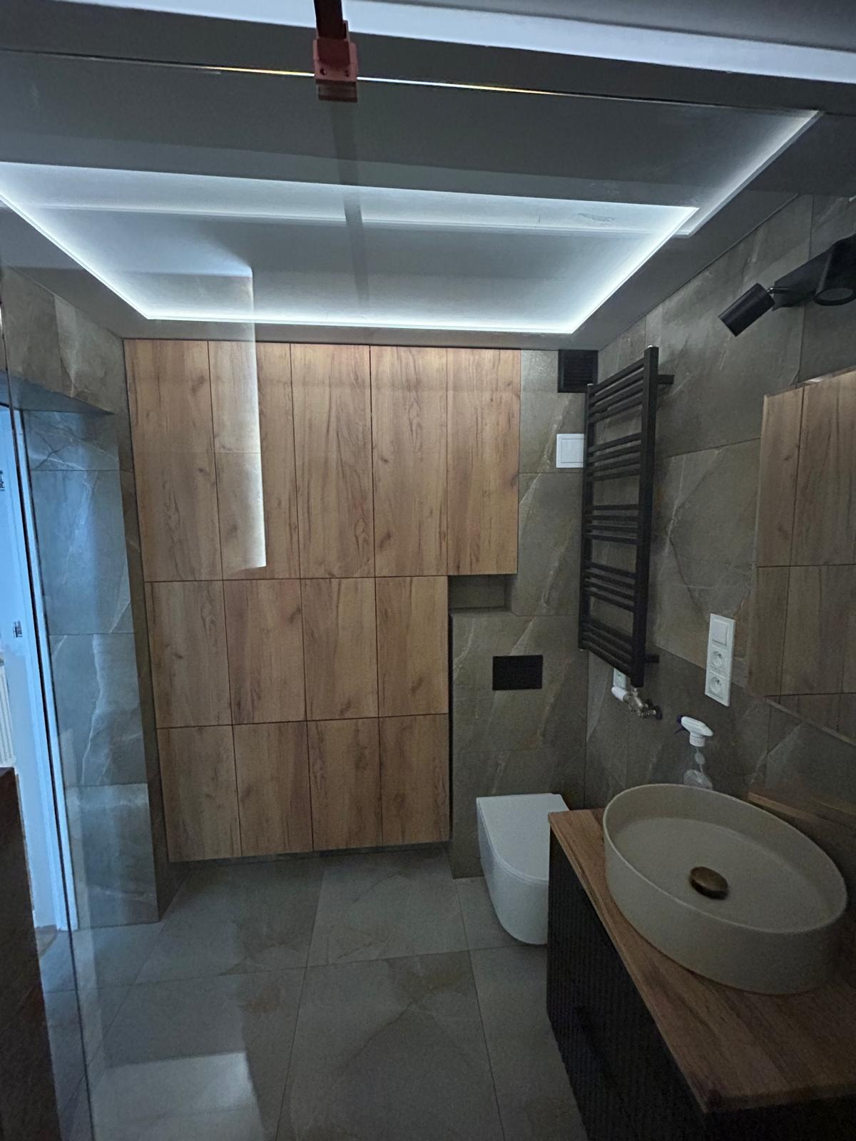 Nowoczesna łazienka z drewnianymi szafkami, szarą armaturą, podświetlanym sufitem i umywalką nablatową. Minimalistyczny design.